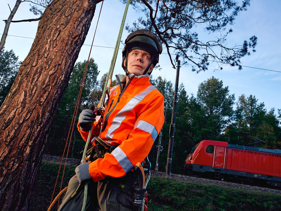 Ein Fahrwegpfleger in Warnkleidung und mit Schutzhelm steht an der Strecke neben einem Baum