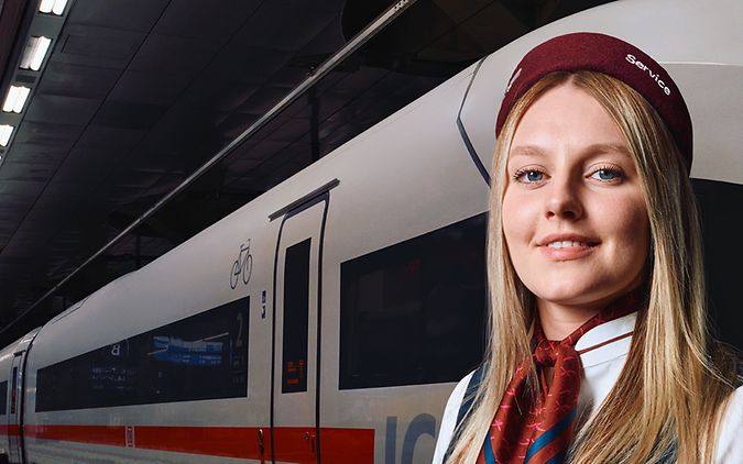 Eine Mitarbeiterin des Bahnhofsservices steht am Bahnsteig eines Bahnhofs, im Hintergrund steht ein ICE im Gleis