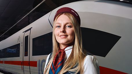 Eine Mitarbeiterin des Bahnhofsservices steht am Bahnsteig eines Bahnhofs, im Hintergrund steht ein ICE im Gleis