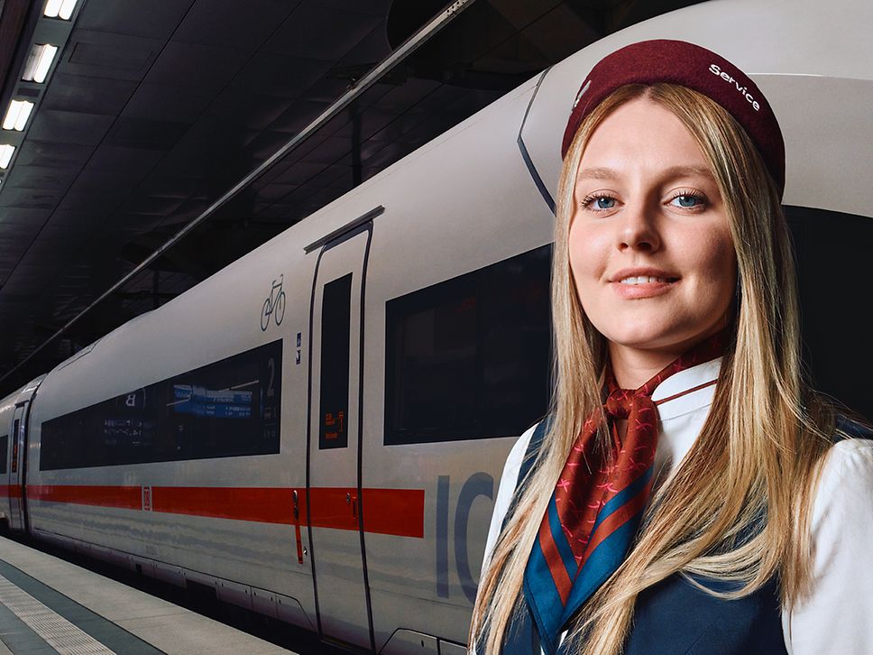 Eine Mitarbeiterin des Bahnhofsservices steht am Bahnsteig eines Bahnhofs, im Hintergrund steht ein ICE im Gleis