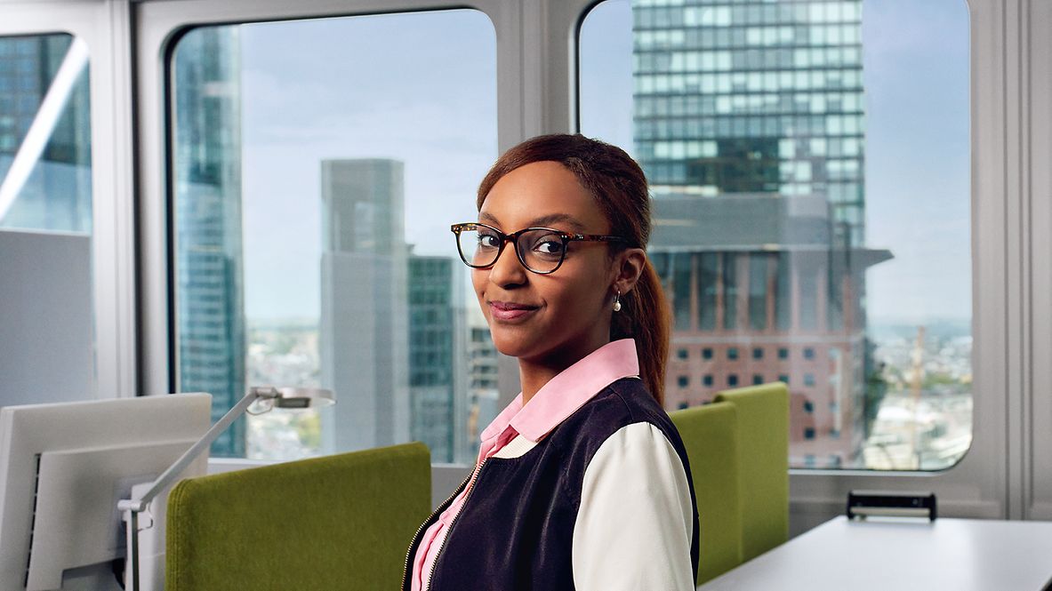 Eine Agility Masterin in Bluse steht in einem Besprechungsraum, im Hintergrund sind Bürogebäude zu sehen