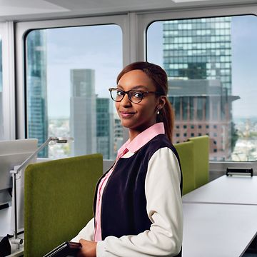 Eine Agility Masterin in Bluse steht in einem Besprechungsraum, im Hintergrund sind Bürogebäude zu sehen