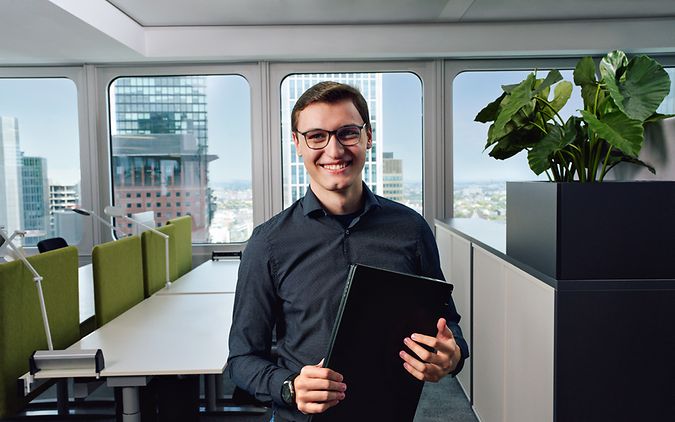 Ein Mann steht lächelnd in einem Großraumbüro und hält einen Laptop vor sich.