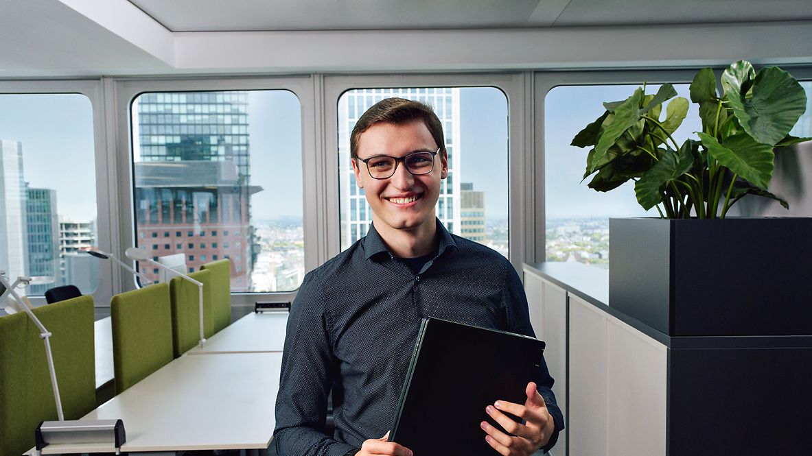 Ein Auszubildender Fachinformatiker für Anwendungsentwicklung steht vor Tischen in einem Büro und hält einen Laptop in der Hand