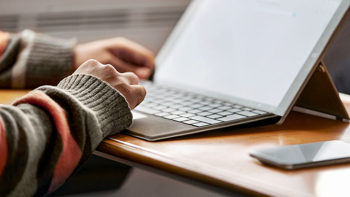 Eine Person sitzt vor einem Schreibtisch mit einem aufgestellten IPad. Daneben liegt ein Smartphone