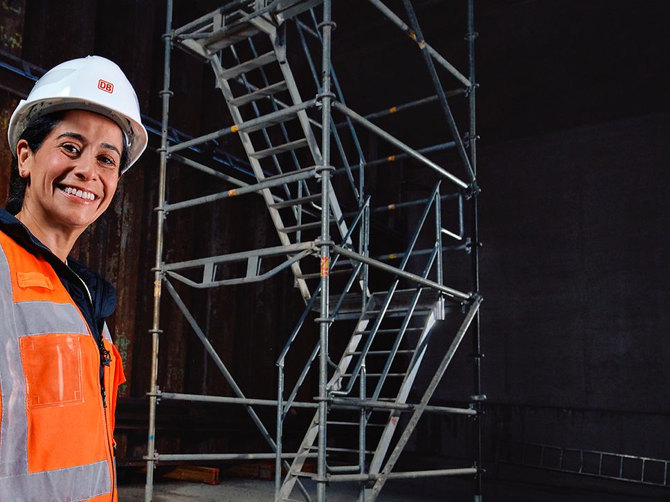 Eine Bauüberwacherin in Warnkleidung und mit Schutzhelm steht vor einer Baustelle