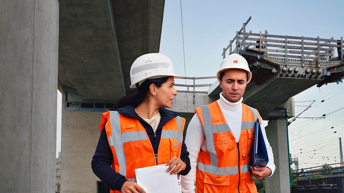 Zwei Bauüberwacher in Warnkleidung und mit Schutzhelm laufen über eine Baustelle und unterhalten sich
