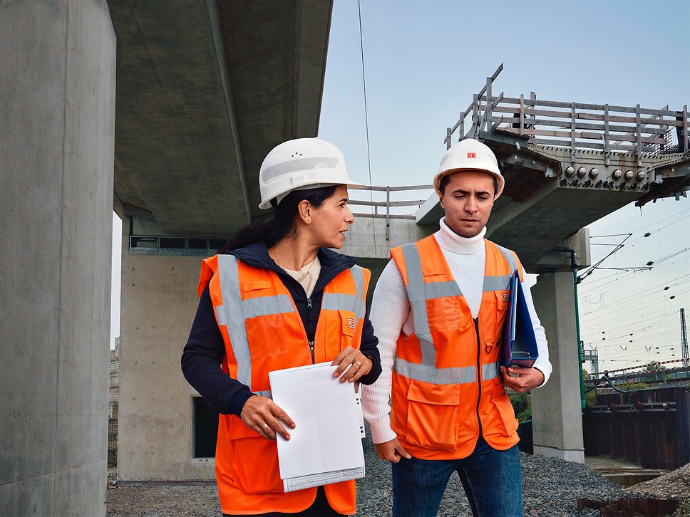 Zwei Bauüberwacher in Warnkleidung und mit Schutzhelm laufen über eine Baustelle und unterhalten sich