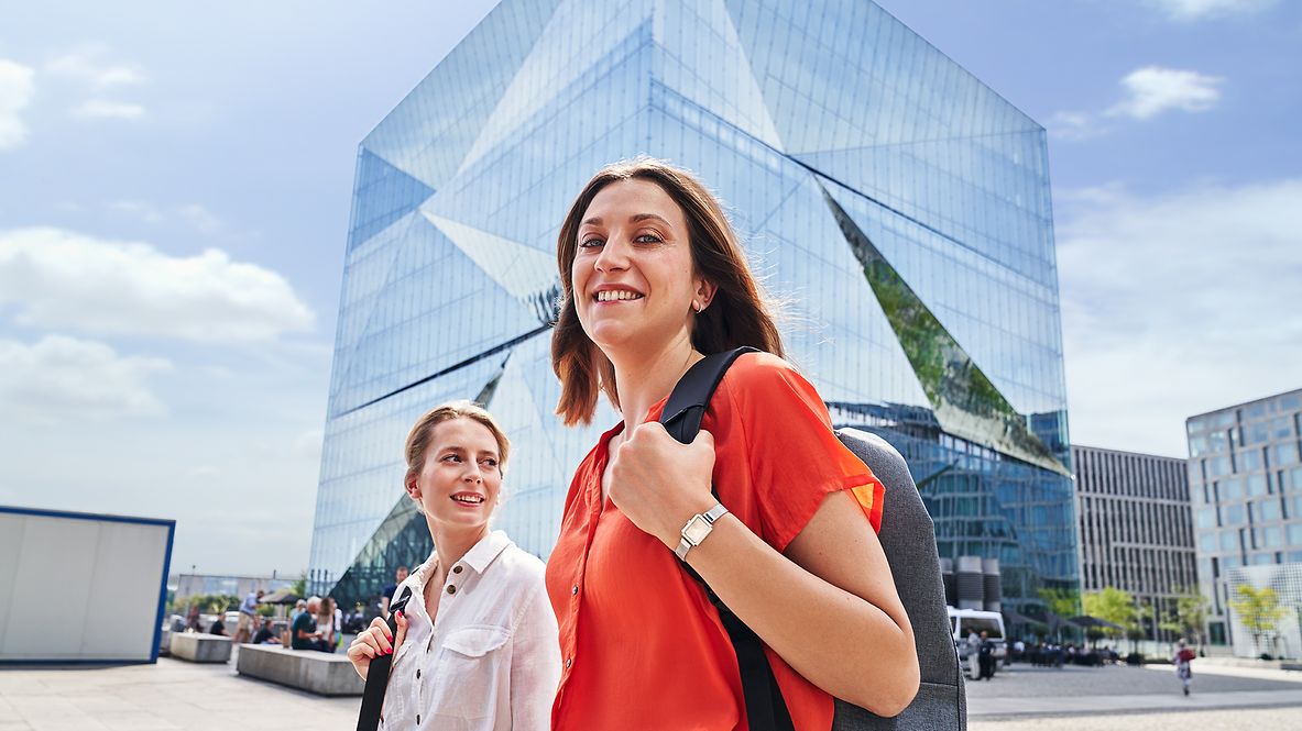 Zwei kaufmännische Mitarbeiterinnen laufen durch die Stadt, im Hintergrund ist ein modernes Bürogebäude zu sehen