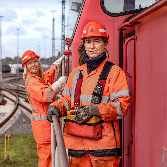 Zwei Lokrangierführerinnen in Warnkleidung und Schutzhelm stehen auf dem Außenpult eines Güterverkehrszuges