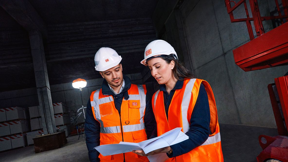 Zwei Ingenieure in Warnkleidung und mit Schutzhelm stehen auf einer Baustelle und sehen sich gemeinsam Planungsunterlagen an