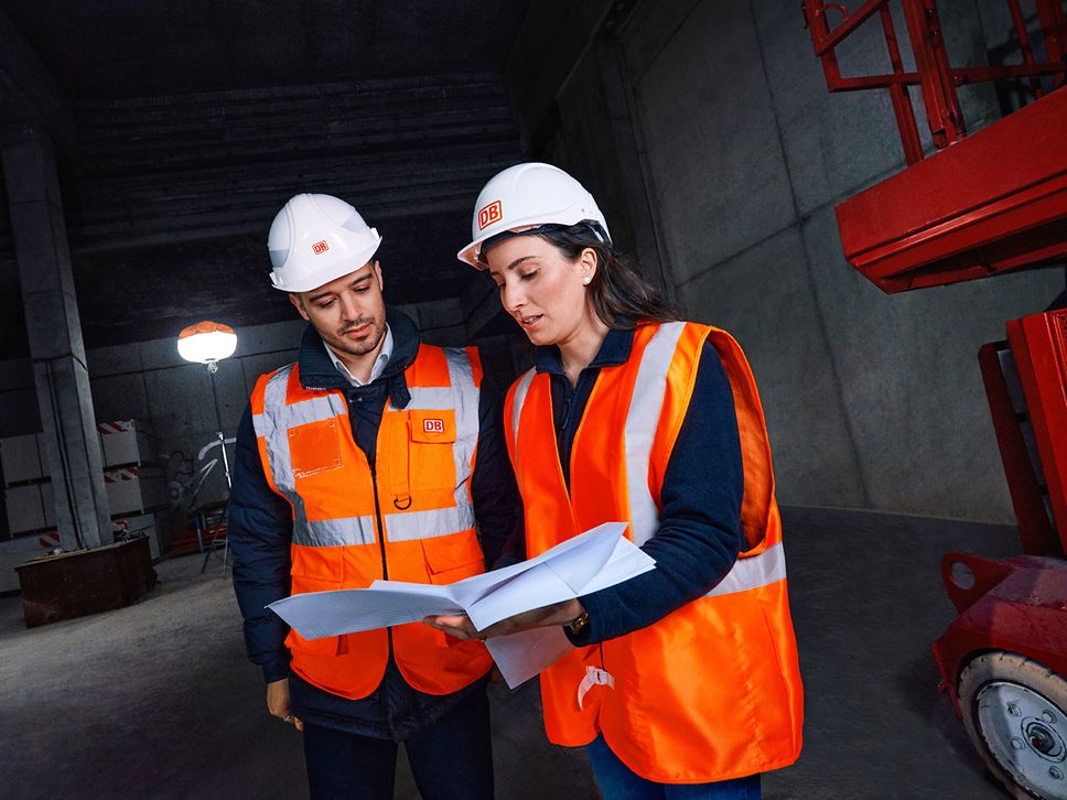 Zwei Ingenieure in Warnkleidung und mit Schutzhelm stehen auf einer Baustelle und sehen sich gemeinsam Planungsunterlagen an