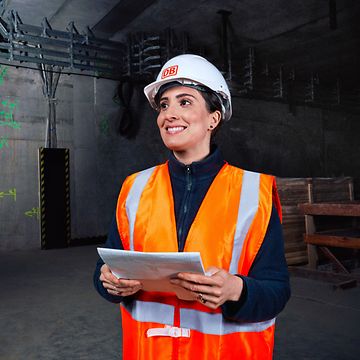 Eine Ingenieurin in Warnkleidung und mit Schutzhelm steht vor einer Baustelle und hält Planungsunterlagen in der Hand