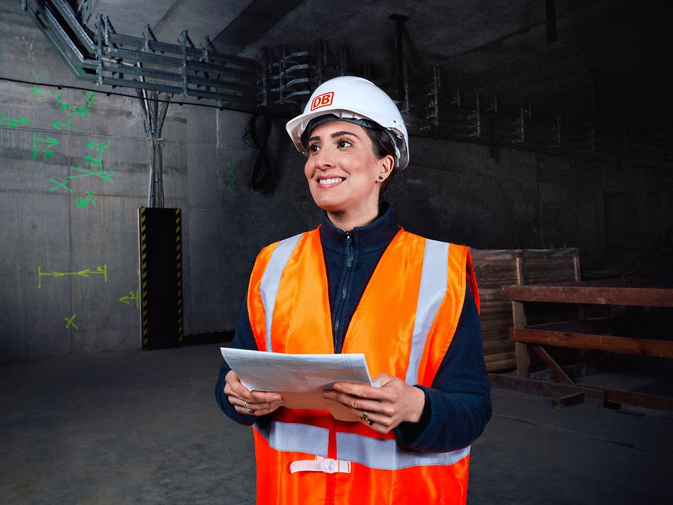 Eine Ingenieurin in Warnkleidung und mit Schutzhelm steht vor einer Baustelle und hält Planungsunterlagen in der Hand