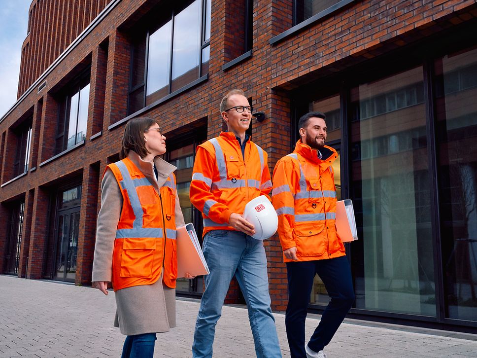 Ein Team von Ingenieuren laufen an einem Gebäude vorbei und halten Schutzhelm und Planungsunterlagen in den Händen