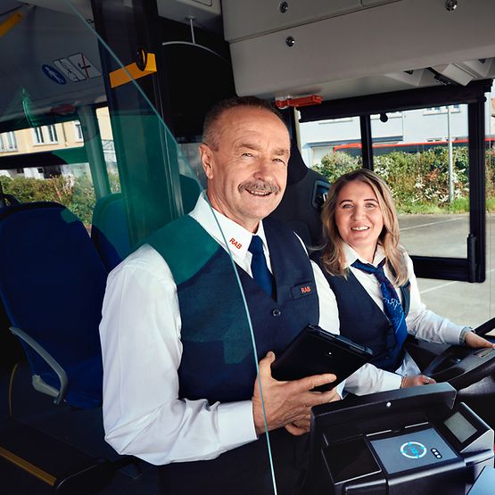 Eine Busfahrerin in Unternehmensbekleidung sitzt am Steuer eines Busses, neben ihr steht ein Busfahrer und hält ein Tablet