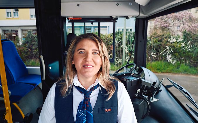Eine Busfahrerin steht in Unternehmensbekleidung vor dem Steuer eines Busses