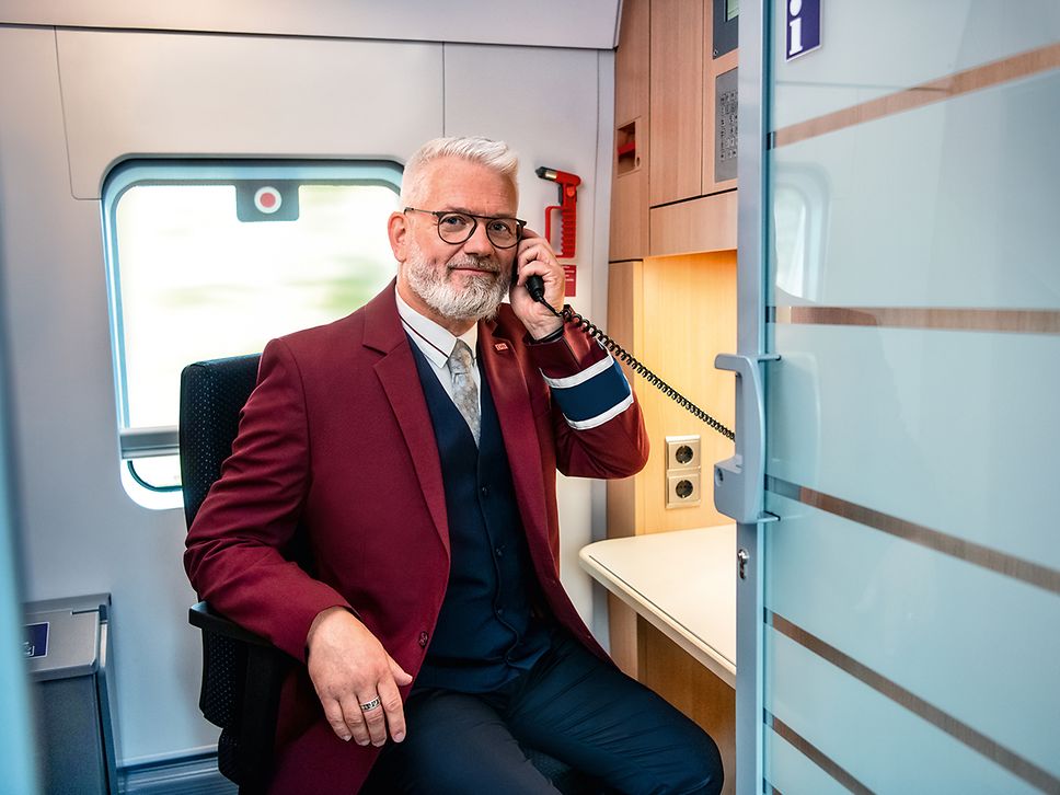 Ein Zugchef in Unternehmensbekleidung sitzt im Zugbegleiter Abteil und telefoniert