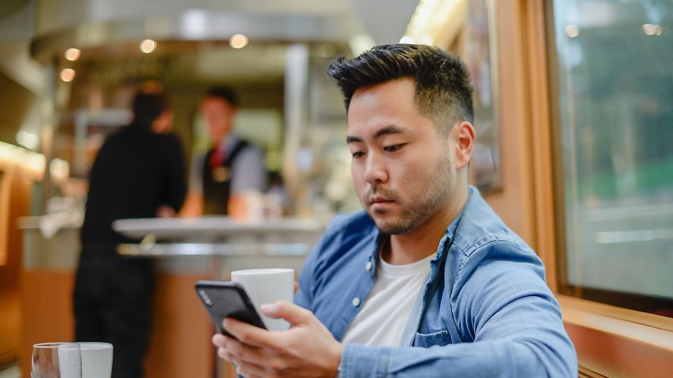 Ein Mann sitzt mit seinem Smartphone und einer Kaffeetasse in der Hand im Bordbistro eines Fernzuges