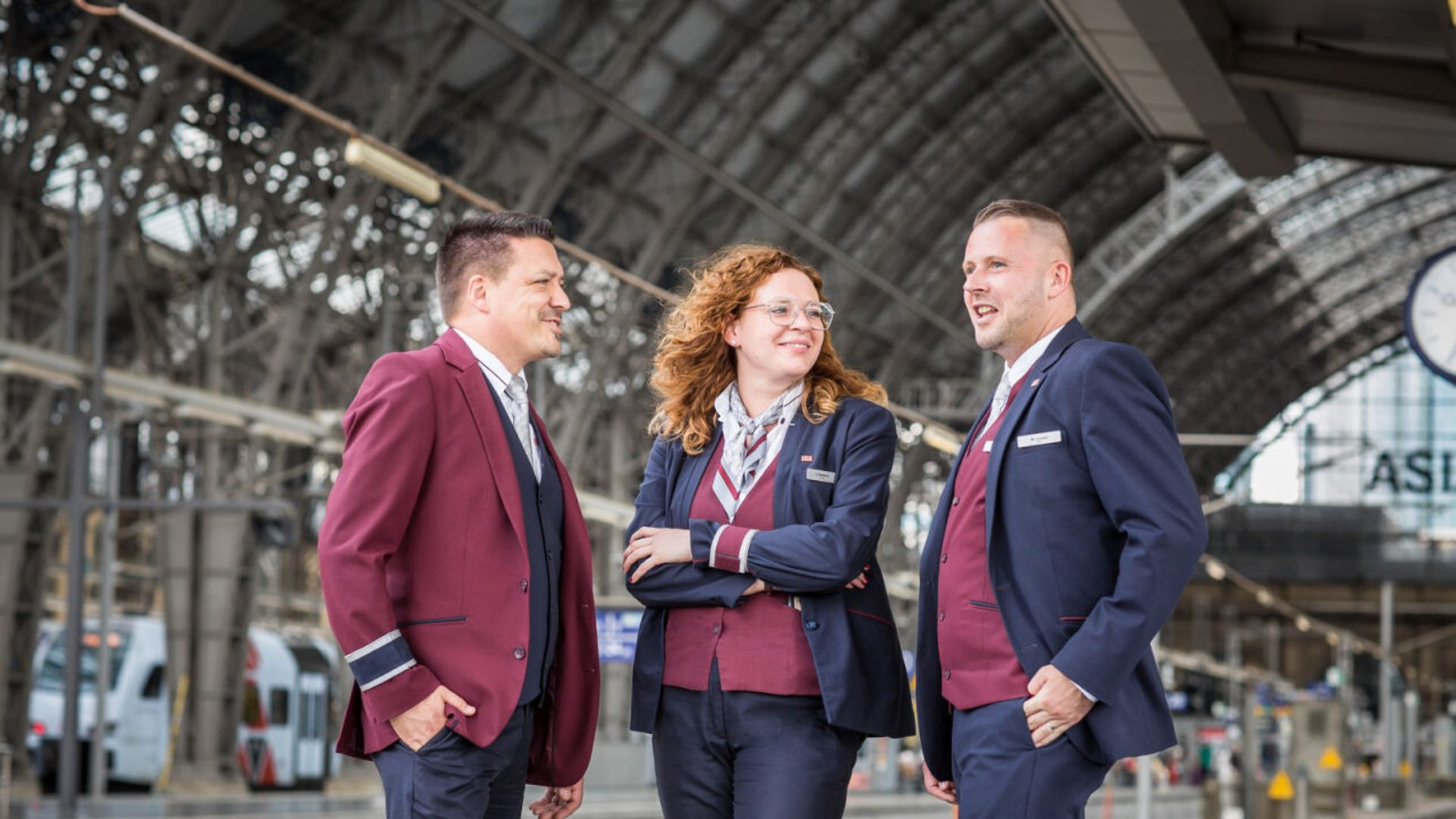 Eine Zugchefin und zwei Zugchefs in Unternehmensbekleidung stehen auf einem Bahnsteig und unterhalten sich