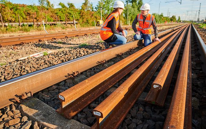 Zwei Gleisarbeiter in Arbeitskleidung knien neben gelagerten Schienen