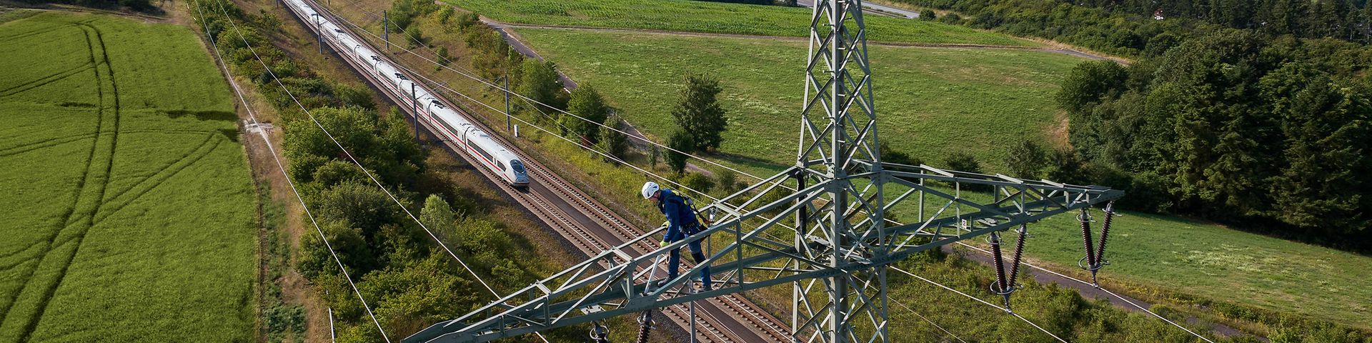 Ein Elektromeister arbeitet auf einer Bahnstromleitung neben einem einem Gleis mit einem heranfahrenden ICE