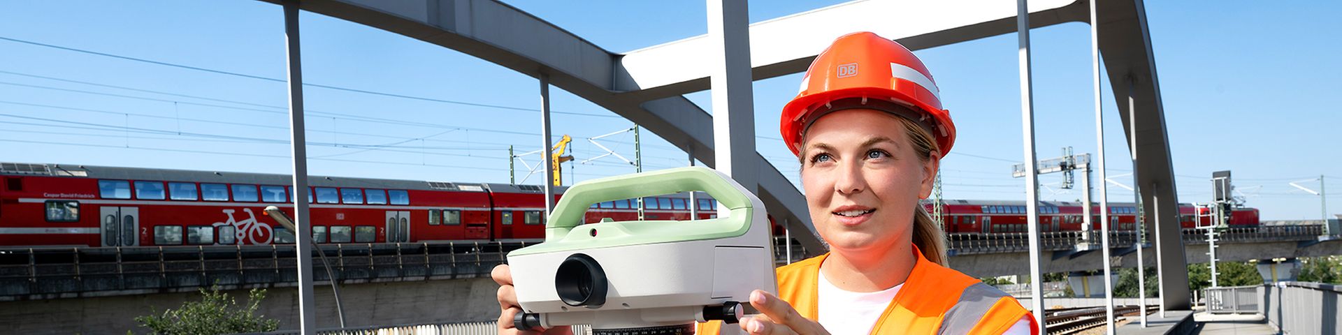 Eine Vermessungsingenieurin in Warnkleidung steht mit einem Tachymeter auf einer Bahnüberführung