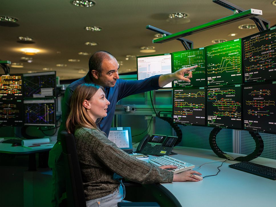 Eine Auszubildende zum Zugverkehrssteuerer sitzt vor den Bildschirmen im Elektronischen Stellwerk neben ihrem Ausbilder