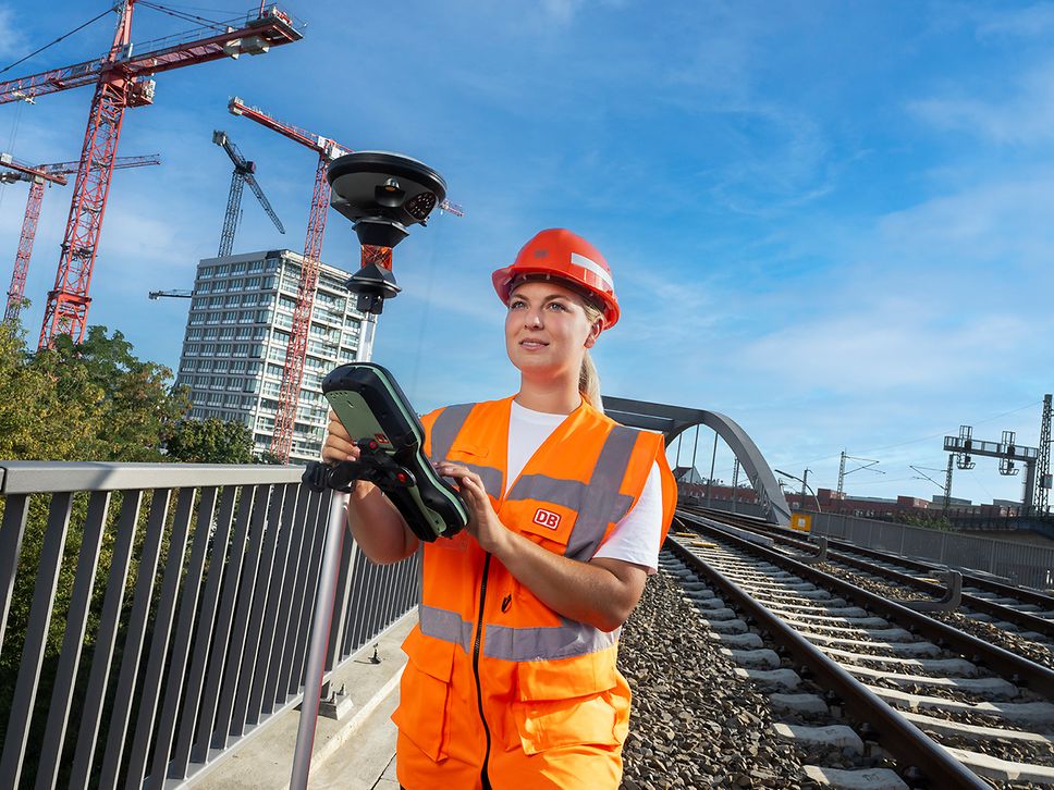 Eine Vermessungsingenieurin läuft in Warnkleidung mit einem GPS-Vermessungsgerät in der Hand auf einer Bahnüberführung