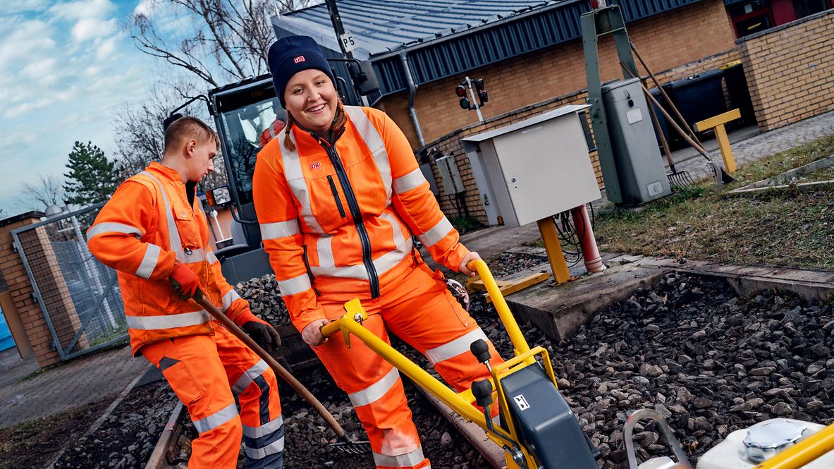 Zwei Auszubildende zum Gleisbauer arbeiten in Schutzkleidung mit Gleisschrauber und Schotterrechen