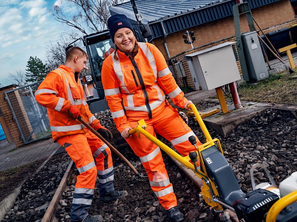 Zwei Auszubildende zum Gleisbauer arbeiten in Schutzkleidung mit Gleisschrauber und Schotterrechen