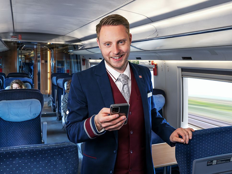 Ein Zugchef in Arbeitskleidung steht im Gang eines fahrenden ICE und hält ein Dienst-Smartphone