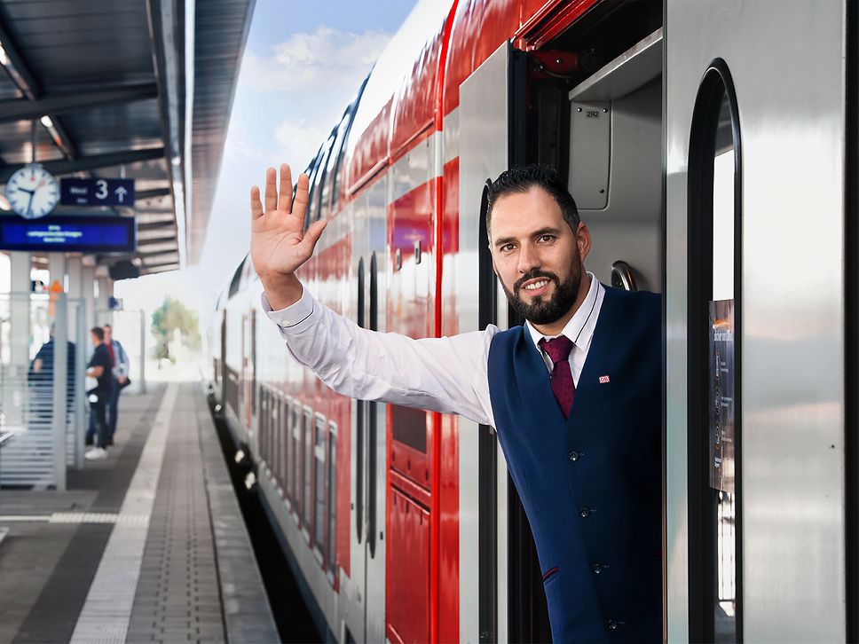 Ein Zugbegleiter lehnt sich aus der Tür eines am Gleis wartenden Regionalzuges und hebt die Hand