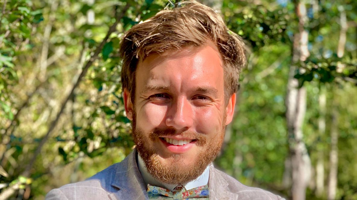 Porträtfoto von Timo Simon, trägt einen beigen Anzug, steht lächelnd in der Natur, hat die Arme verschränkt