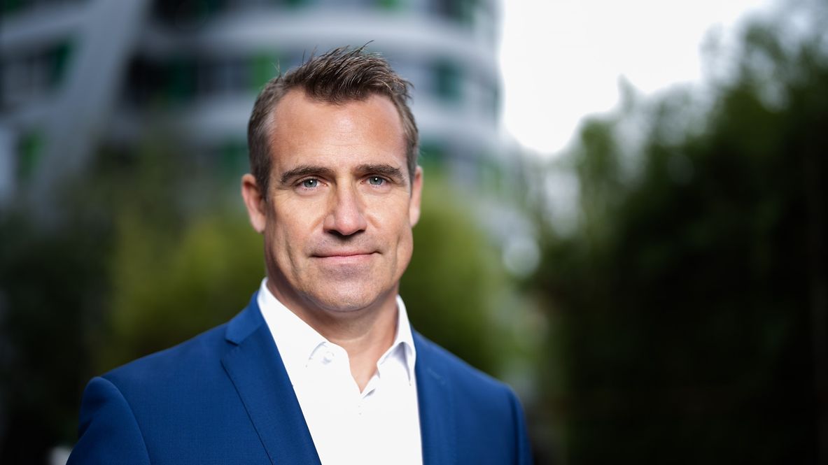 Porträtfoto von Nico Warbanoff, trägt einen blauen Anzug, im Hintergrund sind Bäume und ein Gebäude 