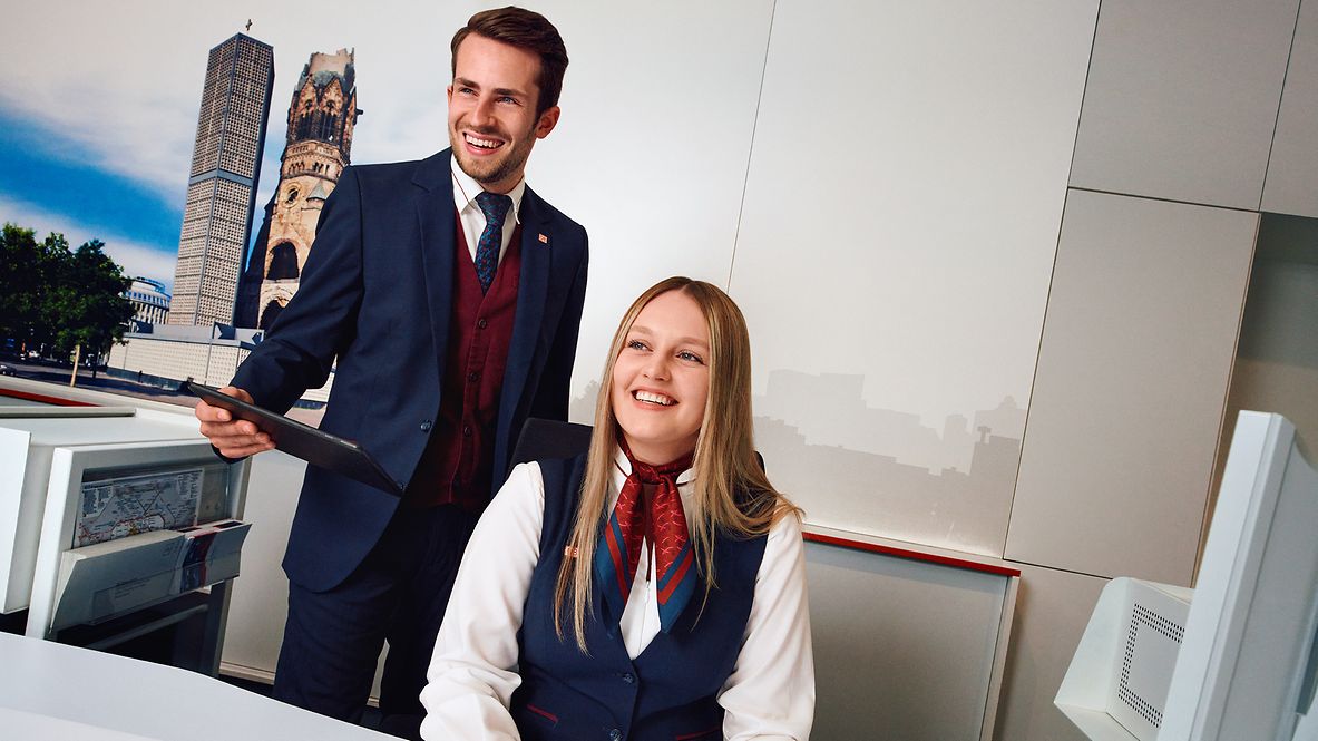 Eine Mitarbeiterin des Bahnhofservices sitzt am Schalter, hinter Ihr steht ein Arbeitskollege und hält ein Tablet in der Hand