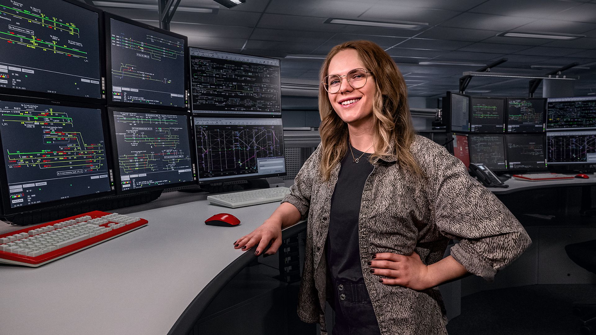 Eine Zugverkehrssteuer:in steht am Schreibtisch vor acht Bildschirmen in einem elektronischen Stellwerk