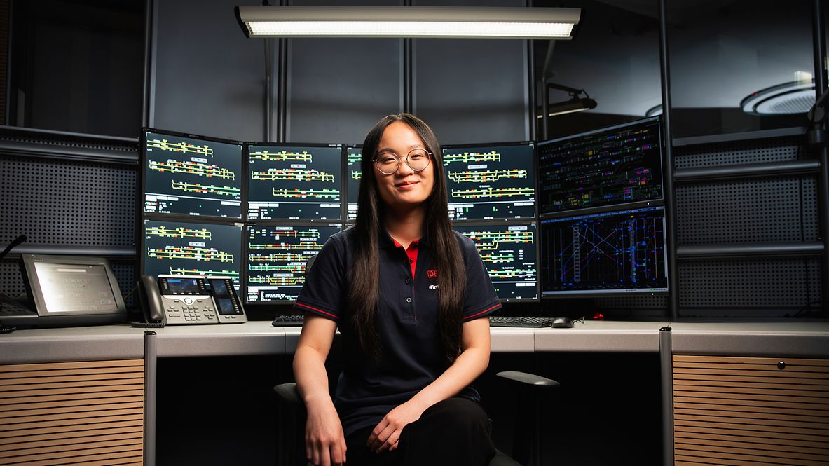 Azubi Zugverkehrssteuerer sitzt mit dem Rücken an ihrem Arbeitsplatz vor zehn Monitoren