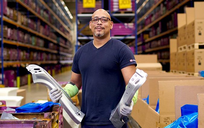 A male warehouse clerk working for DB Schenker is standing in a warehouse holding two components in his hands