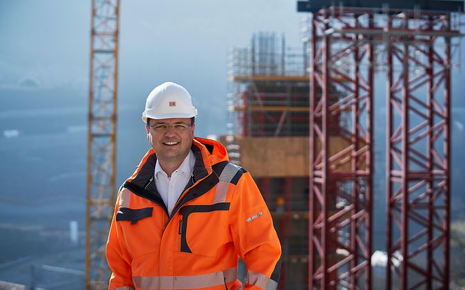 Bauingenieur Jörg steht mit orangener Sicherheitsweste und Helm vor seiner Baustelle. Im Hintergrund steht ein großer Kran.