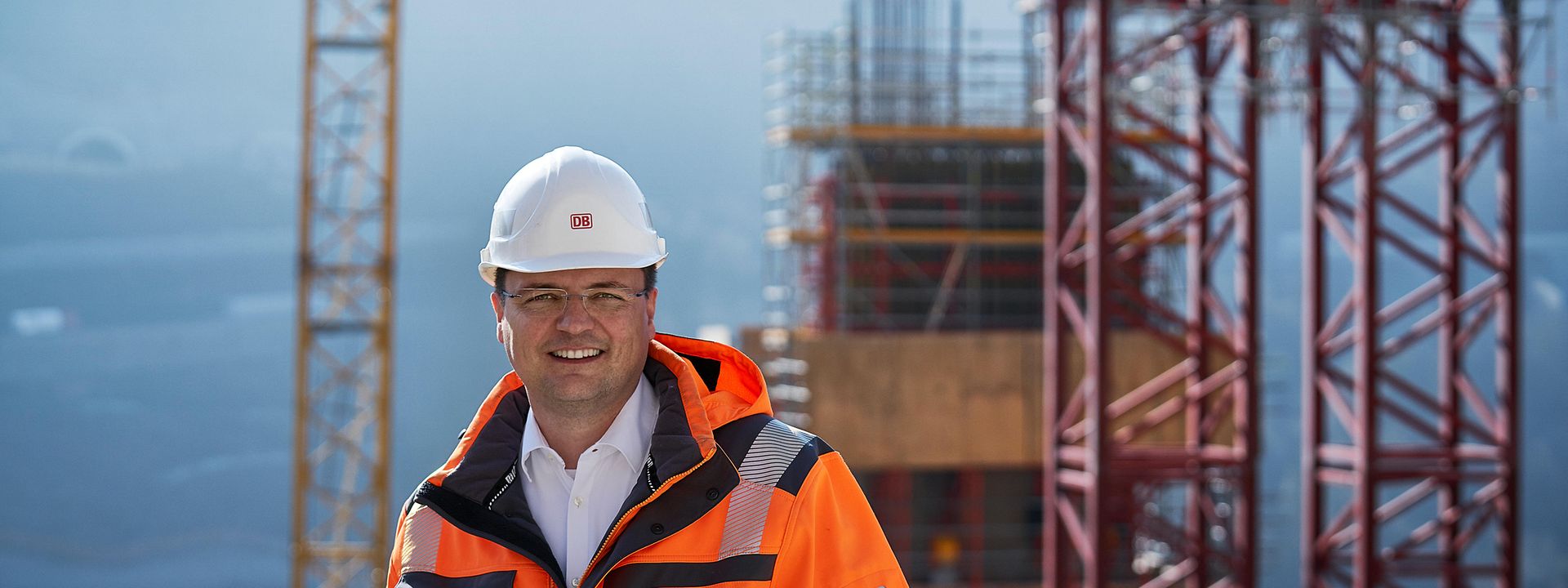 Bauingenieur Jörg steht mit orangener Sicherheitsweste und Helm vor seiner Baustelle. Im Hintergrund steht ein großer Kran.