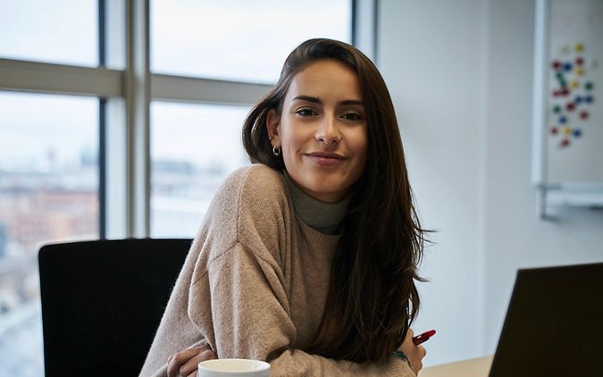 Eine junge Frau sitzt mit Kaffe zufrieden am Laptop im Büro.