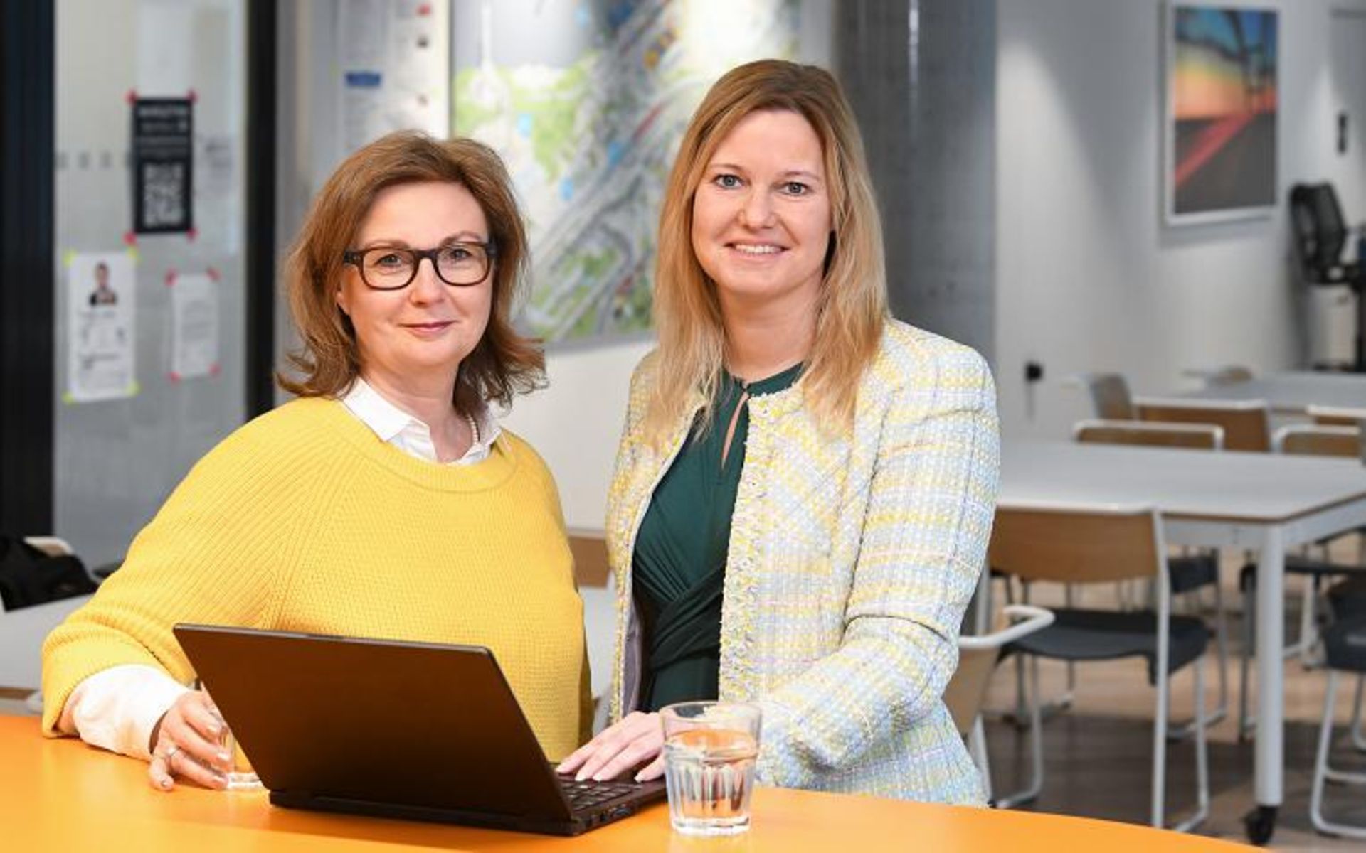 Die Leiterinnen Personalmanagement Carola und Catherine stehen lächelnd im Großraumbüro nebeneinander vor einem Laptop.