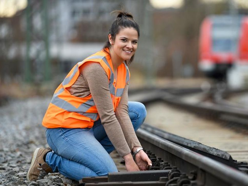 Die junge Fahrdienstleiterin Vanessa arbeitet bei der DB Netz AG. Sie kniet mit Schutzweste am Gleisbett und lächelt.