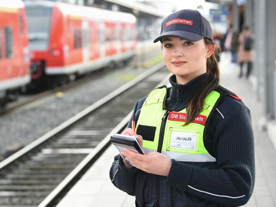 Quelle: Deutsche Bahn AG Die junge Fachkraft für Schutz und Sicherheit Sofie steht in Dienstkleidung mit Block und Stift ausgestattet auf einem Gleis.