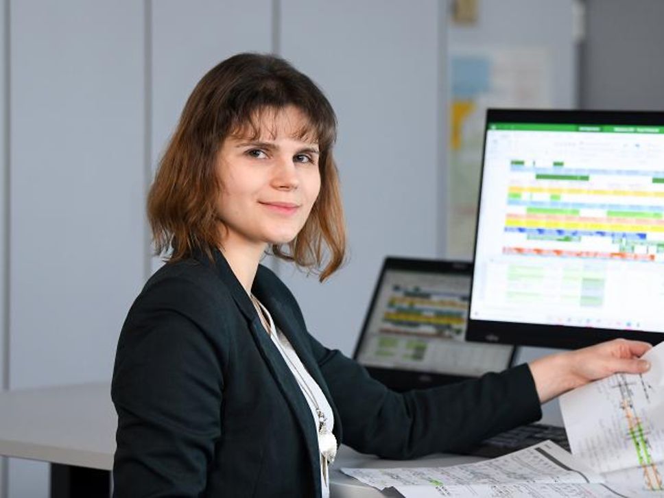 Quelle: Deutsche Bahn AG Bautechnologin Riccarda sitzt im Büro vor ihrem Bildschirm am Schreibtisch und hält lächelnd einen Bauplan.