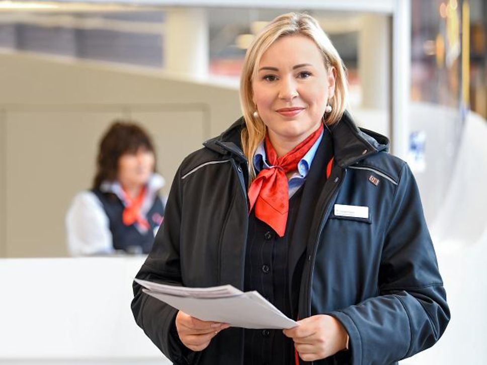 Die Leiterin Bahnhofsservice Regina steht lächelnd in ihrer blauen Uniform mit rotem Halstuch in der Bahnhofshalle.