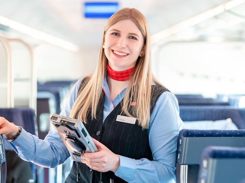 Die junge Zugbegleiterin Ulrike steht im ICE zwischen den Sitzreihen und trägt lächelnd ihre blaue Uniform mit rotem Halstuch.