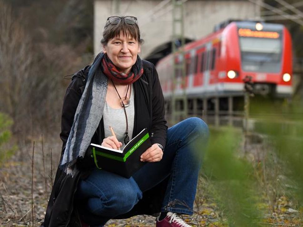 Katja ist Projektingenieurin Umwelt bei DB Netz in Frankfurt am Main. Sie kniet und im Hintergrund fährt ein Regionalzug vorbei.