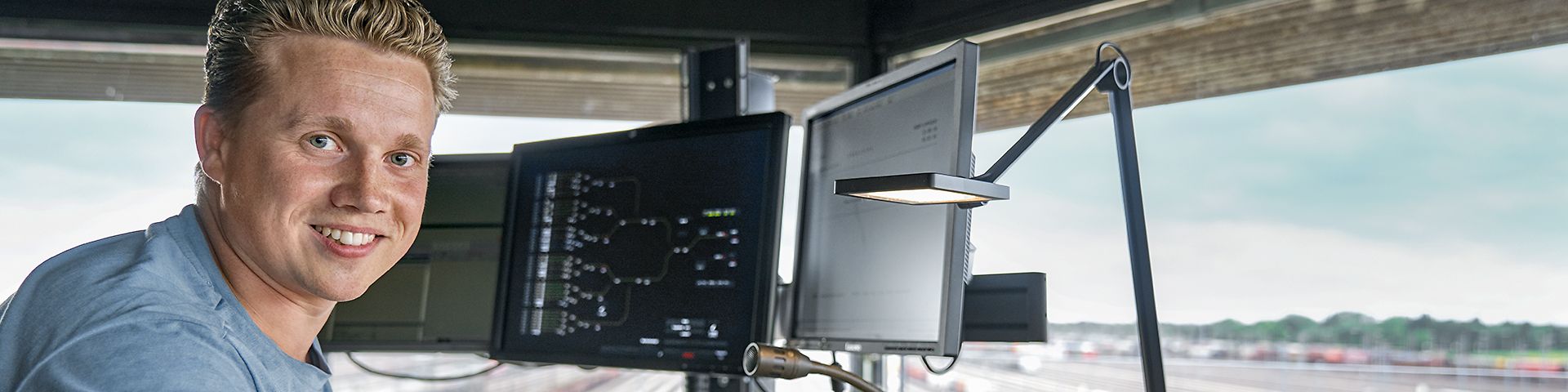 Ein Auszubildender zum Zugverkehrssteuerer steht vor drei Bildschirmen in einem Stellwerk